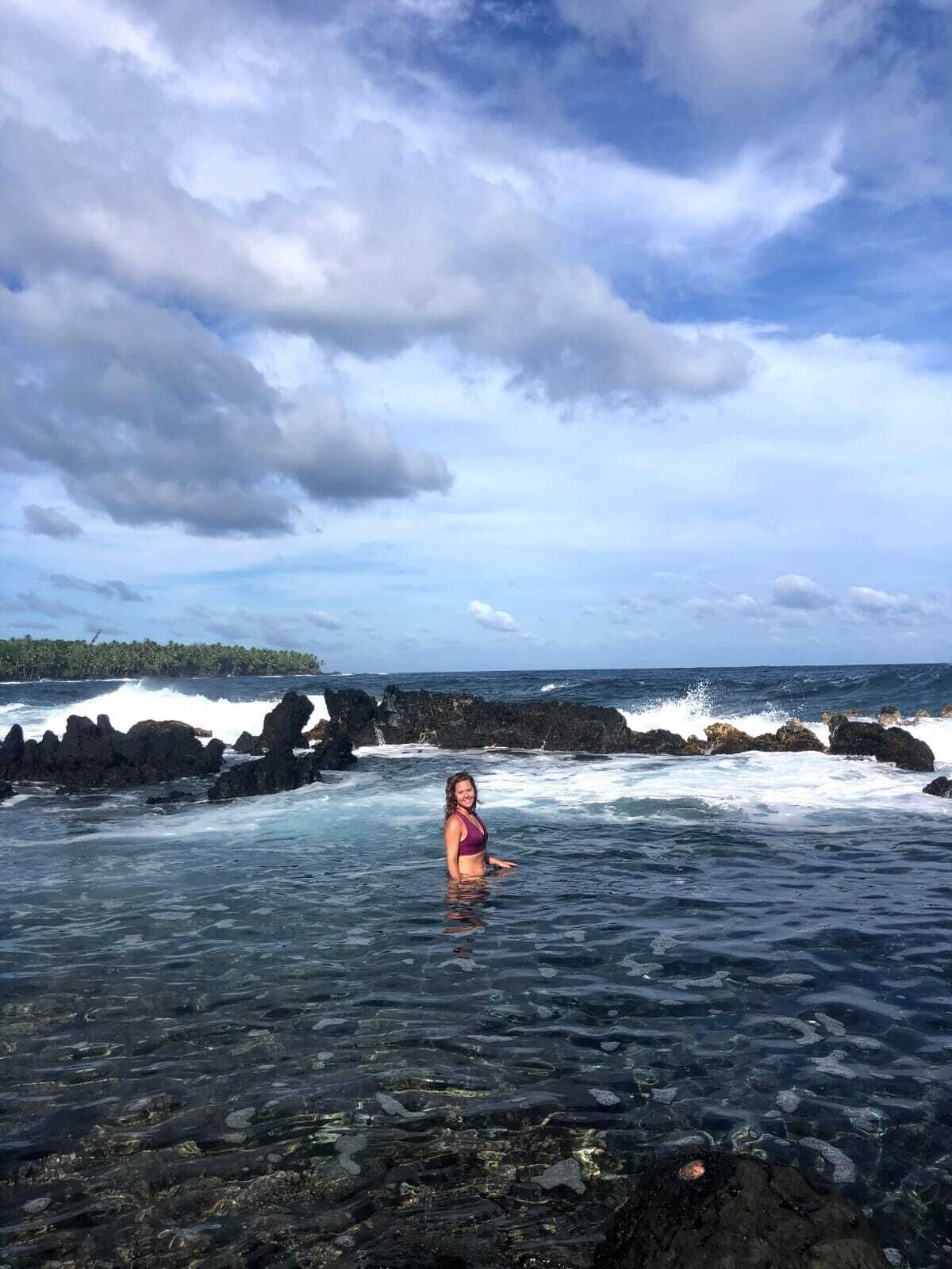 Una persona de pie en aguas cristalinas cerca de formaciones rocosas, con vista al océano y un cielo nublado.