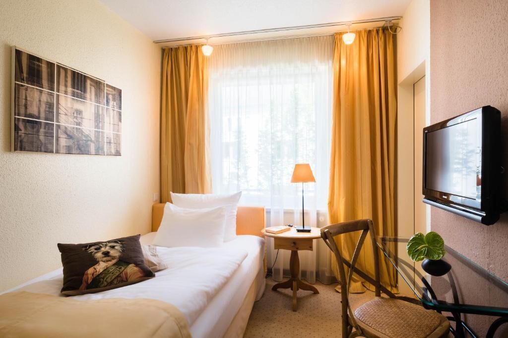A hotel room with a bed, a chair, a small table, a lamp, a TV, and a window with curtains.