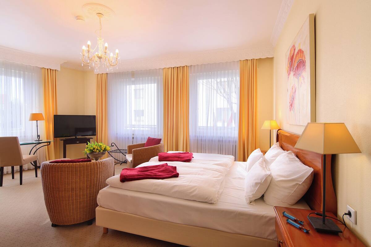 A hotel room with a large bed, a television, a desk with a computer, a wicker chair, and a lamp.