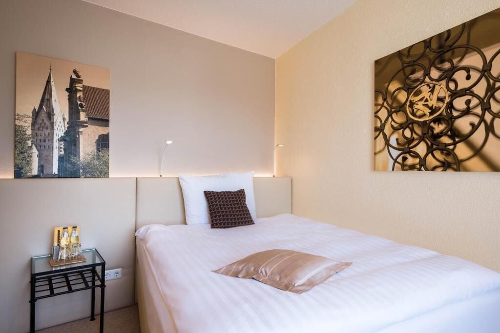 A neatly made bed with a white comforter and a single brown pillow, a small bedside table with a glass top, and a wall-mounted artwork depicting a building.