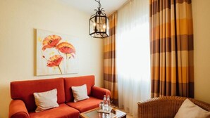 A living room with a red sofa, a wicker chair, a coffee table with water bottles, and a painting of flowers on the wall. - Hotel zur Mühle (Paderborn)