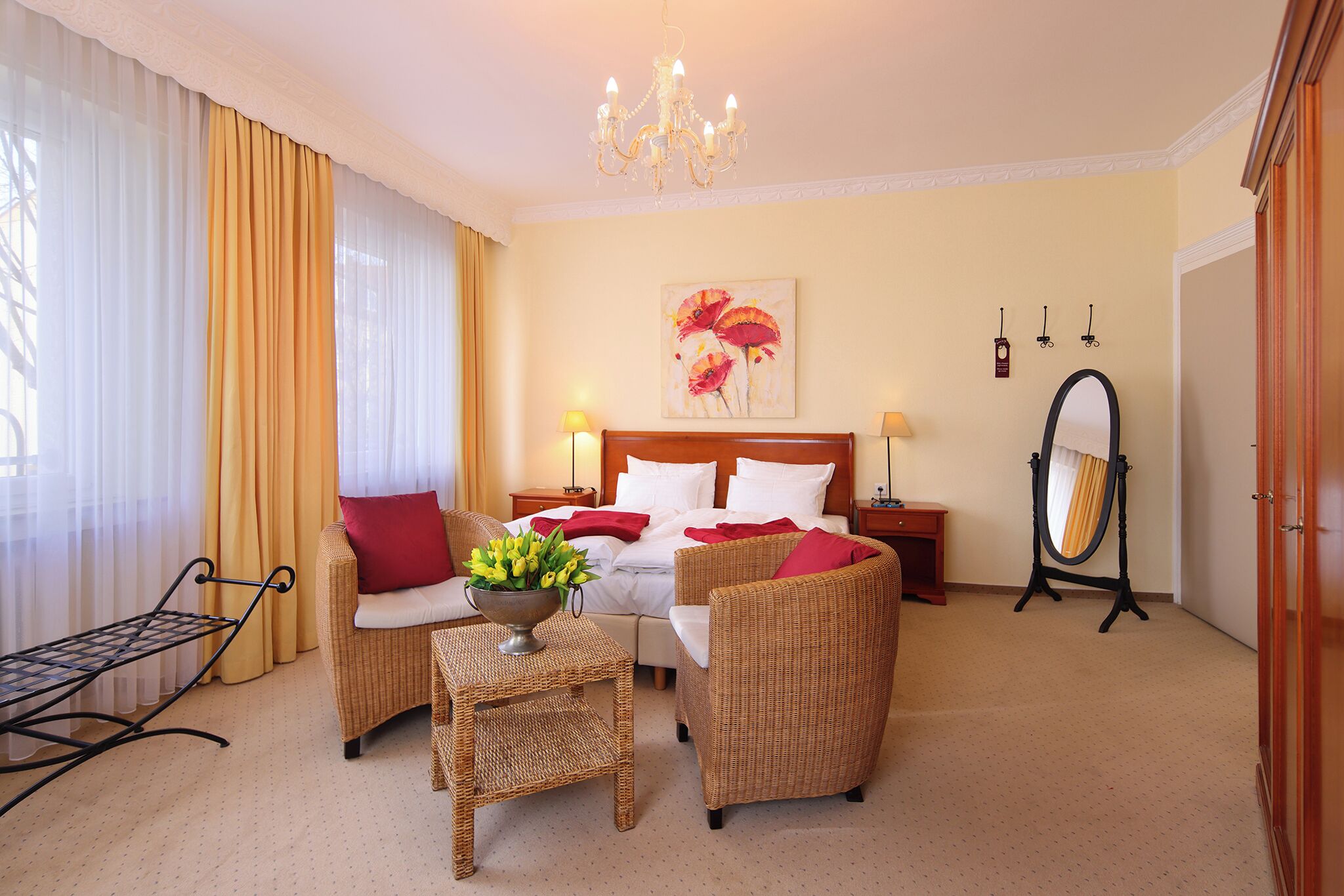 Ein Hotelzimmer mit einem Bett, zwei Korbstühlen, einem kleinen Tisch mit einer Vase, einem Spiegel und einer Chandelier.