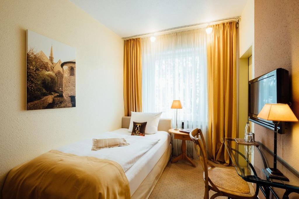 Una habitación de hotel con cama, escritorio, silla, lámpara, televisión y un cuadro en la pared.