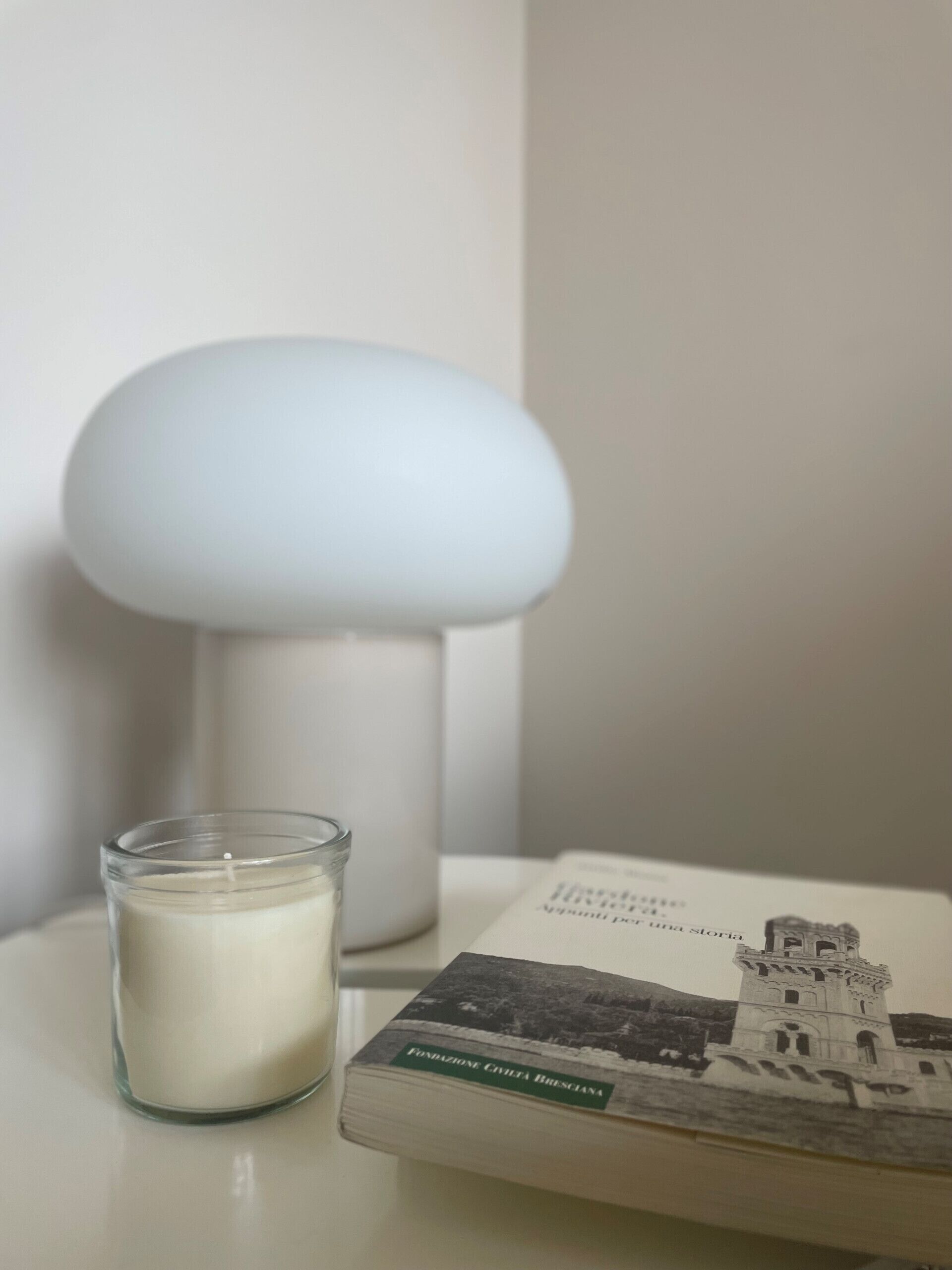 A candle in a glass jar, a book titled 'Fondazione Civiltà Bresciana', and a white lamp on a table.