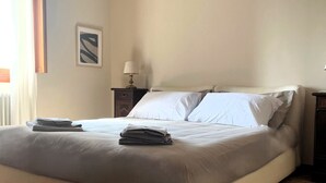 Una cama bien hecha con almohadas y toallas dobladas, una mesita de noche con una lámpara, y un cuadro en la pared.