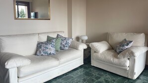 Una habitación con un sofá y un sillón blancos, ambos con cojines estampados, una mesa redonda con lámpara y un espejo en la pared.