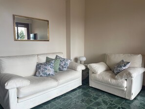 Ein Zimmer mit einem weißen Sofa und einem Sessel, beide mit blumigen Kissen, einem runden Tisch mit einer Lampe und einem Spiegel an der Wand.