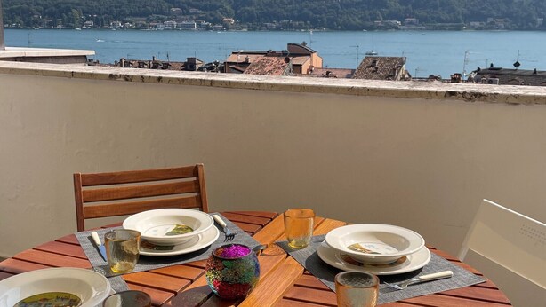 A table set for dining with plates, glasses, and cutlery, overlooking a body of water and a hillside. - Mansarda del Gallo_Penthouse with Lake View and Swimming Pool (Salò)