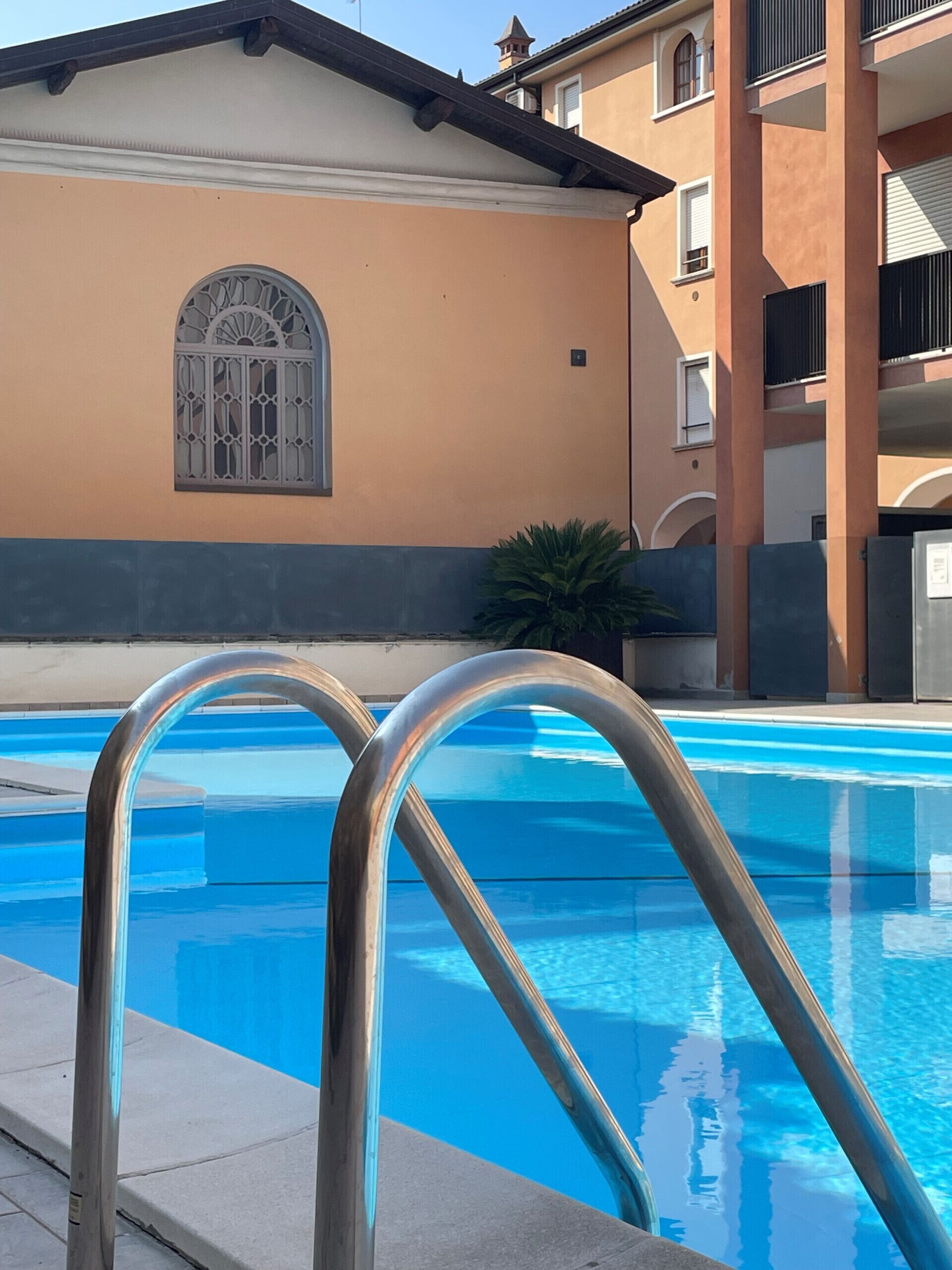 A swimming pool with metal railings, a building with a beige facade and a window with decorative bars, and a plant near the building.