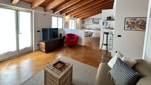 A spacious living area with wooden beams, a television, a red chair, a sofa, and a kitchen in the background. - Mansarda del Gallo_Penthouse with Lake View and Swimming Pool (Salò)