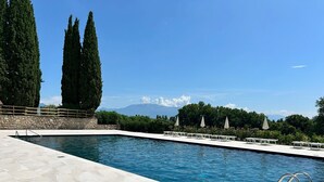 Una piscina rectangular con borde de piedra, rodeada de sillas de descanso y sombrillas blancas, con un muro de piedra y altos árboles al fondo.