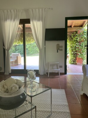 Ein Zimmer mit Glastisch, weißen Vorhängen, Blick ins Freie und Fliesenboden.
