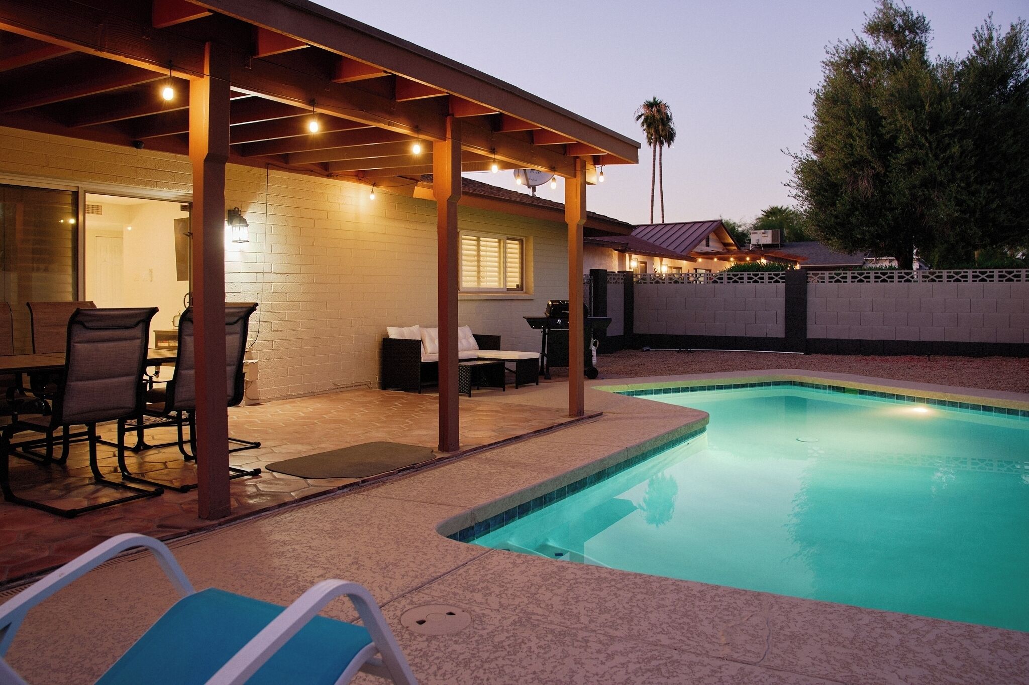 A backyard with a swimming pool, covered patio with chairs, and a barbecue area.