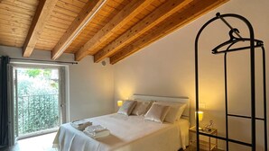 A bedroom with a wooden ceiling, a bed with white linens, a bedside table with a lamp, and a hanging chair. - Borgo Alba Lake View Loft_Design in the Historic Village (Polpenazze del Garda)