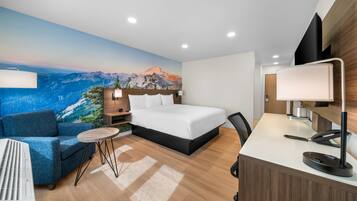 Habitación de hotel con cama, escritorio, silla, sofá, una mesita, lámpara y un cuadro de un paisaje montañoso.