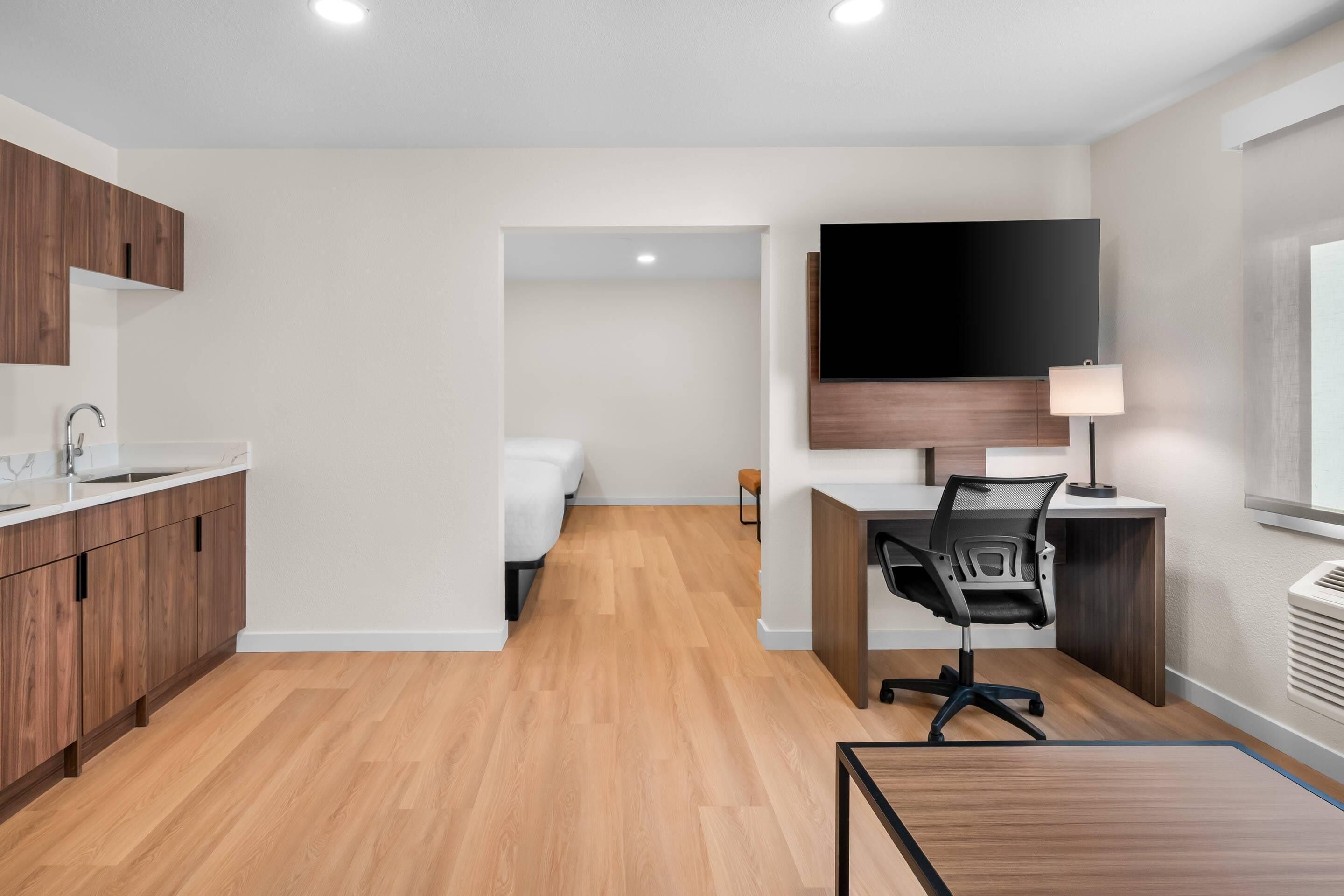 A modern hotel room with a wooden floor, a desk with a chair, a flat-screen TV mounted on the wall, a bedside table, and a window.