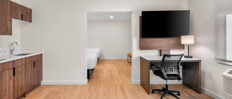 A modern hotel room with a wooden floor, a desk with a chair, a flat-screen TV mounted on the wall, a bedside table, and a window.