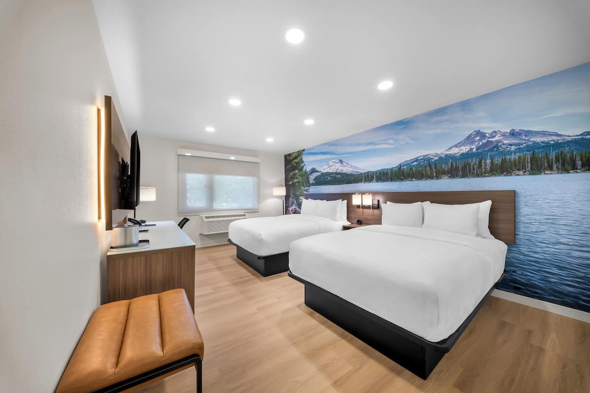 A hotel room with two beds, a desk, a bench, a TV, and a large window.