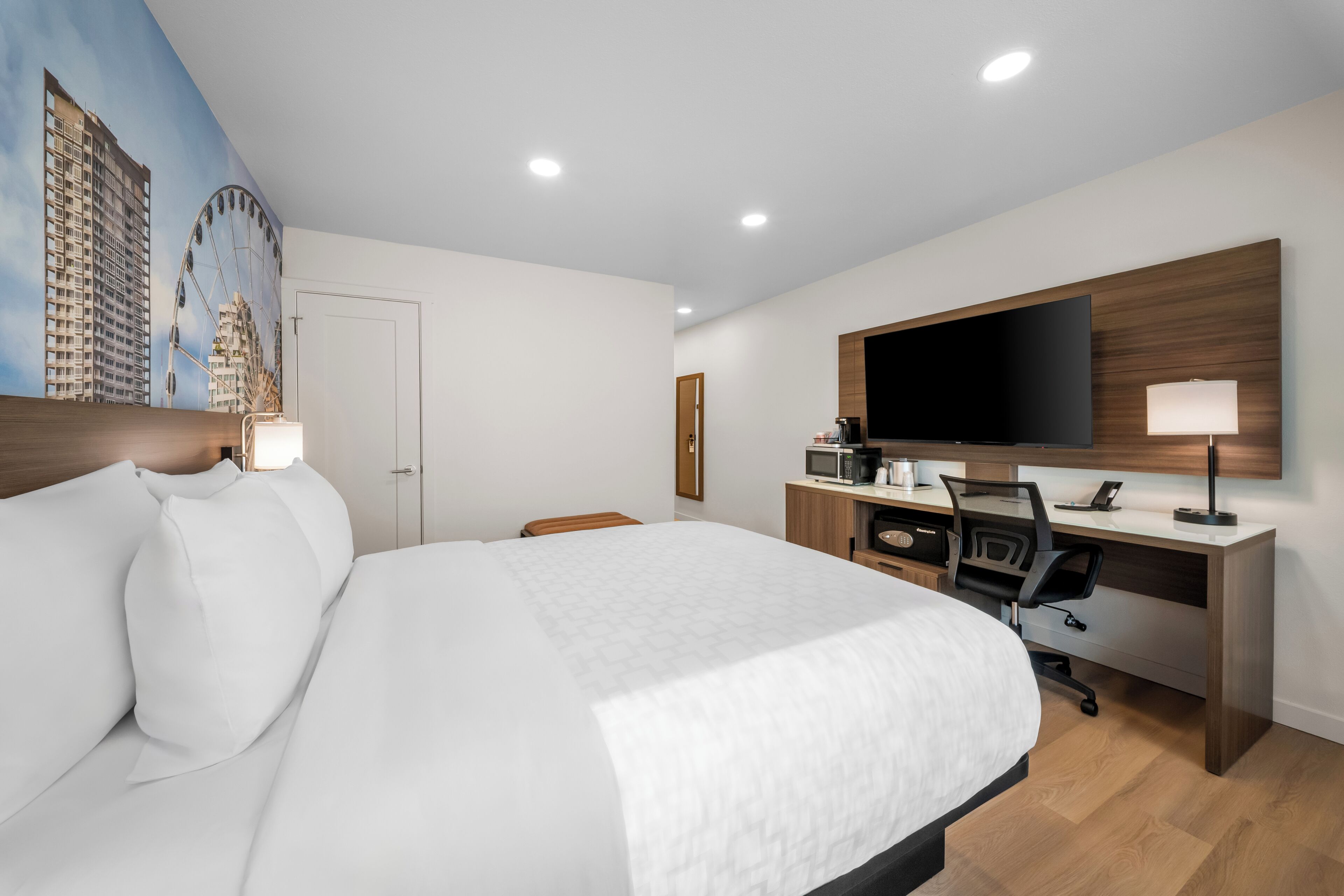 A hotel room with a large bed, a desk with a computer, a flat-screen TV, and a mural of buildings on the wall.