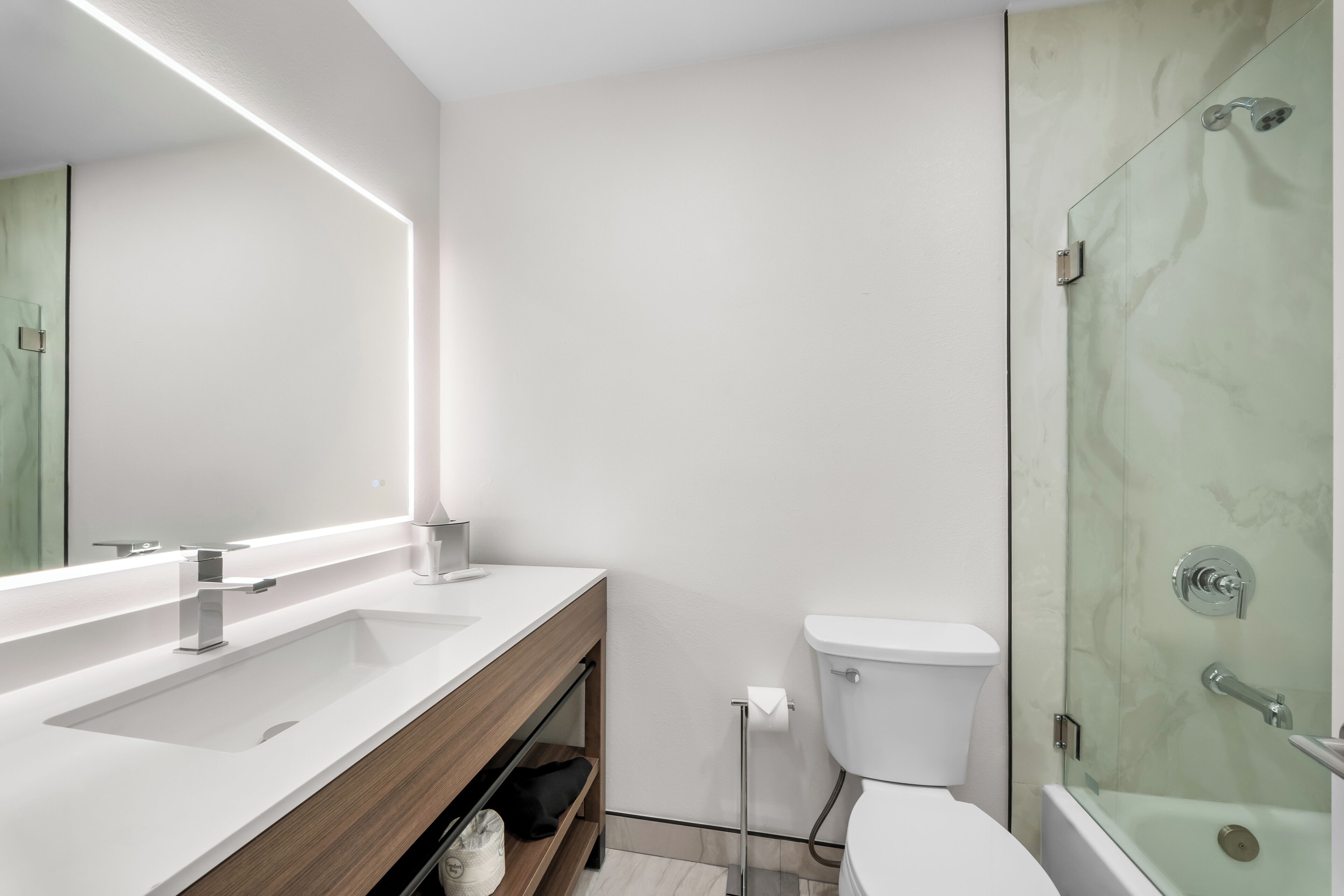 A modern bathroom with a white sink, a mirror with under-cabinet lighting, a toilet, and a glass-enclosed shower.