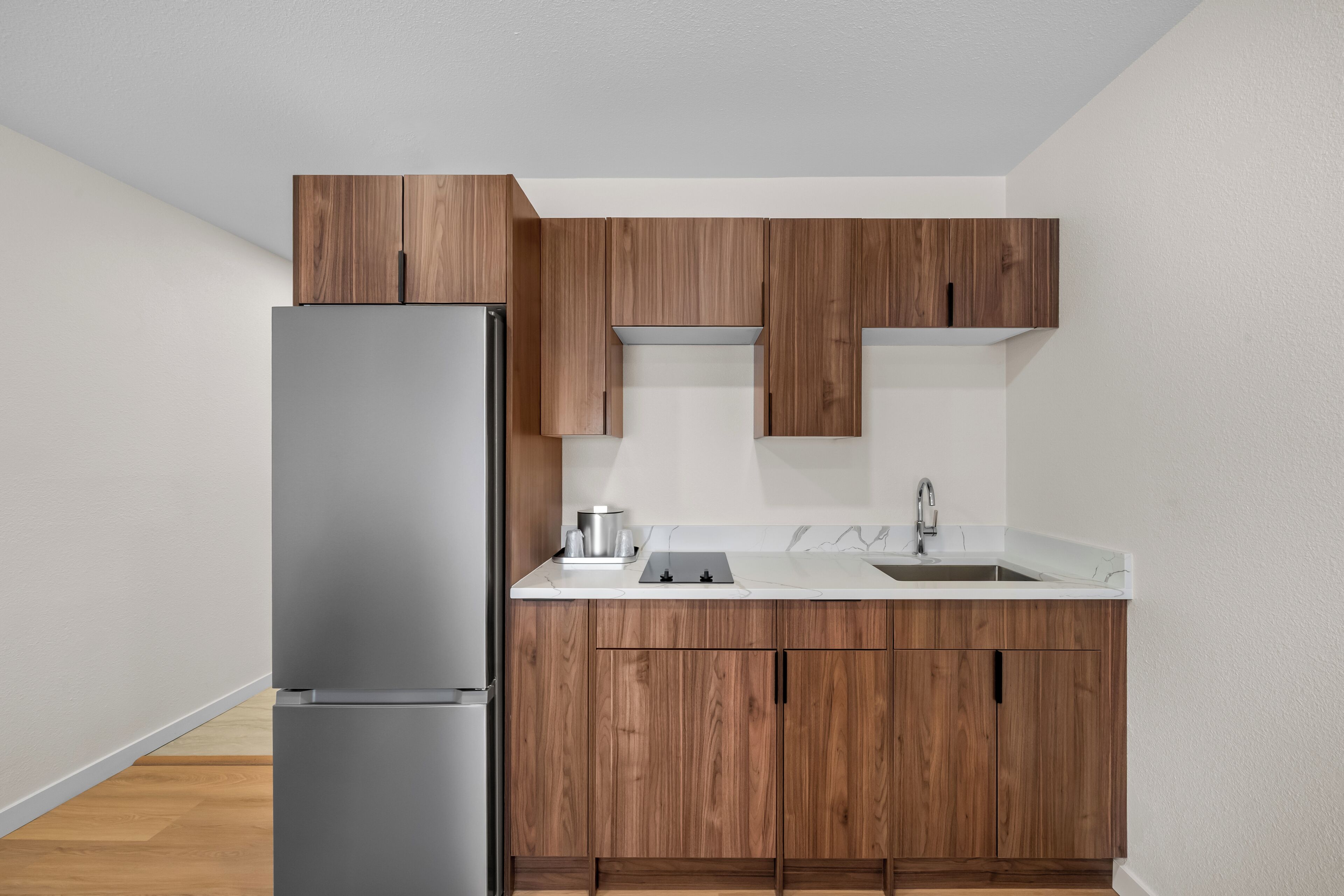 A kitchen with wooden cabinets, a stainless steel refrigerator, a white countertop, and a sink.