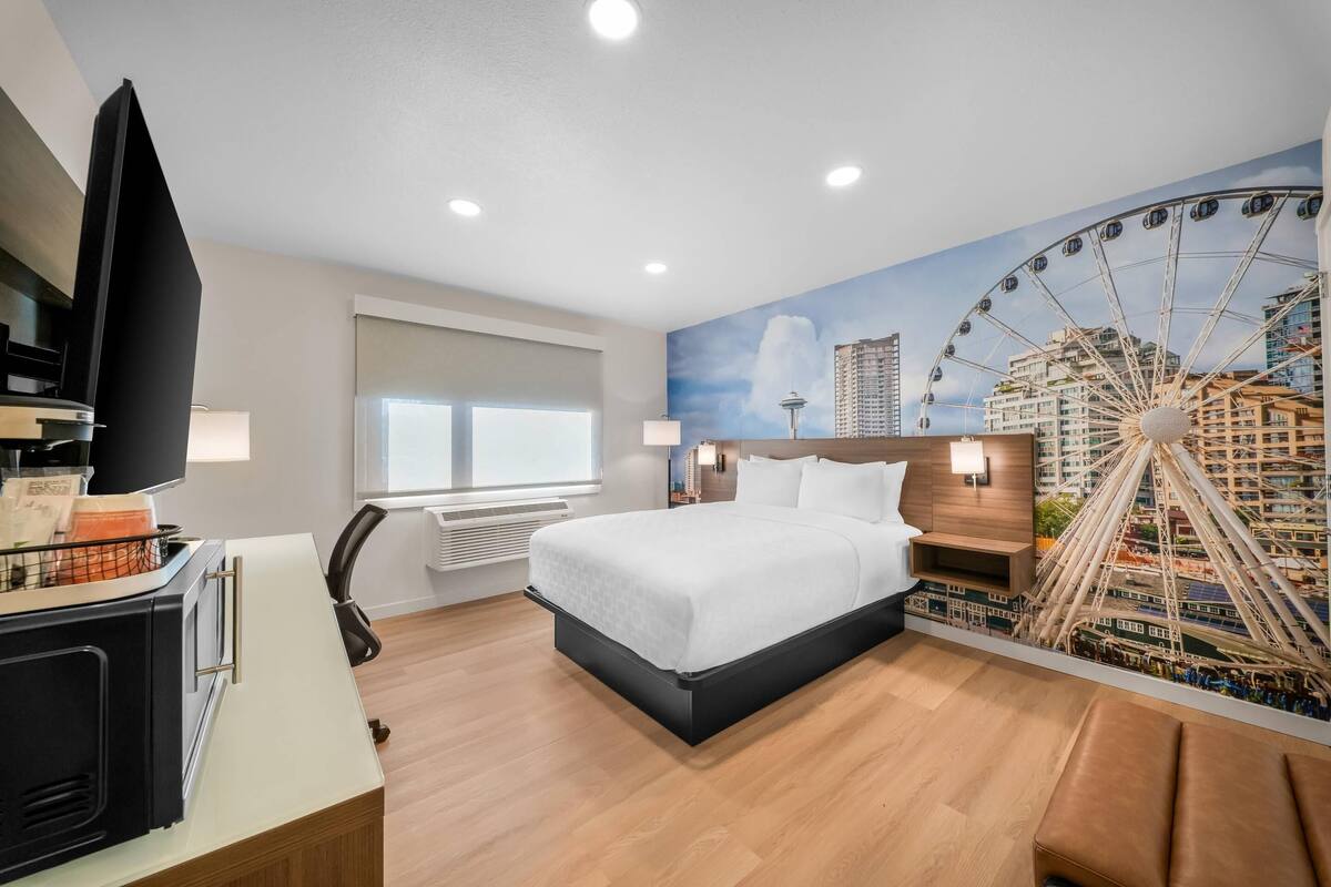 A hotel room with a bed, a TV, a desk, a chair, a window, and a mural of a Ferris wheel.