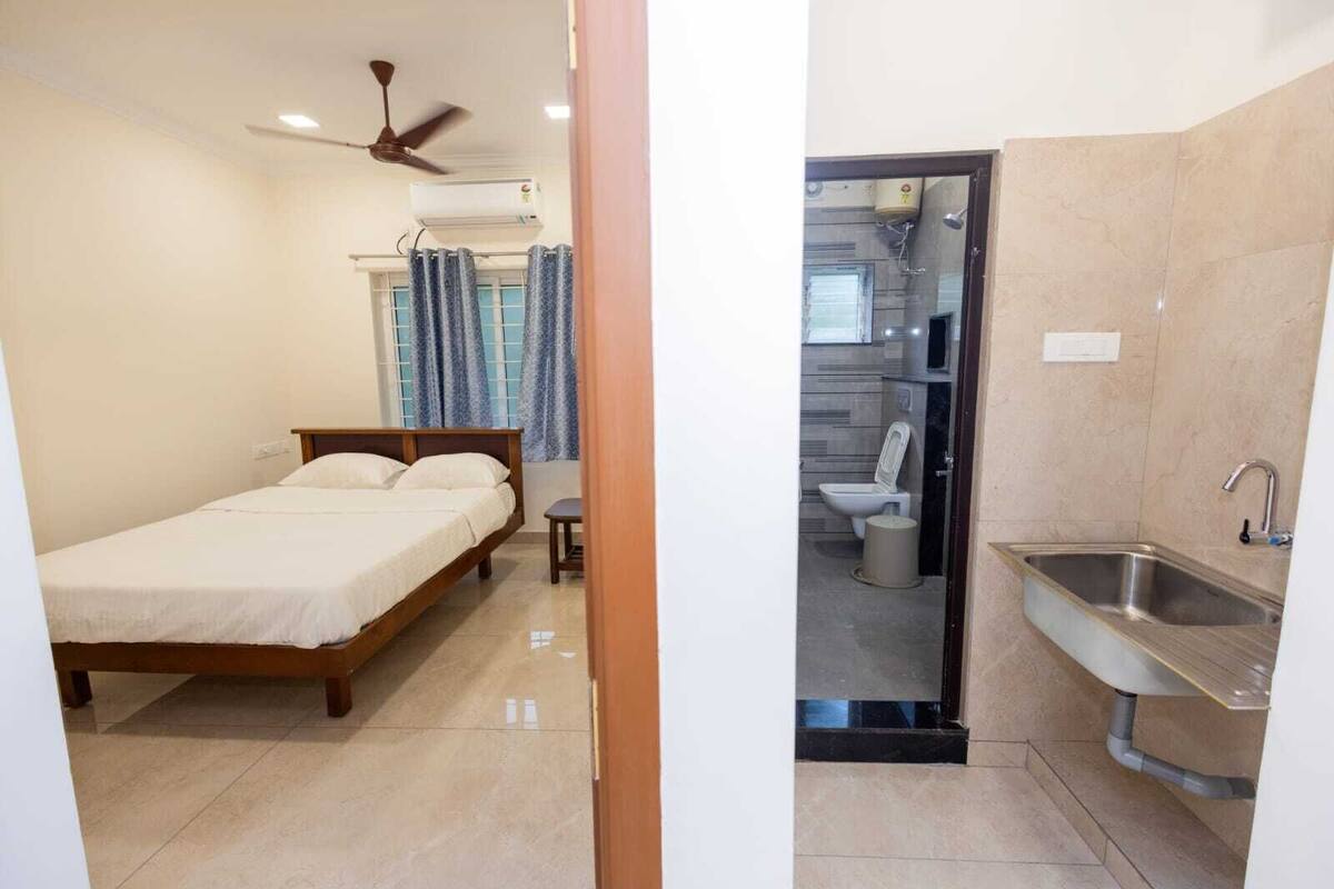 A bedroom with a bed, a ceiling fan, and a window with curtains. The adjacent bathroom includes a toilet, sink, and shower area.