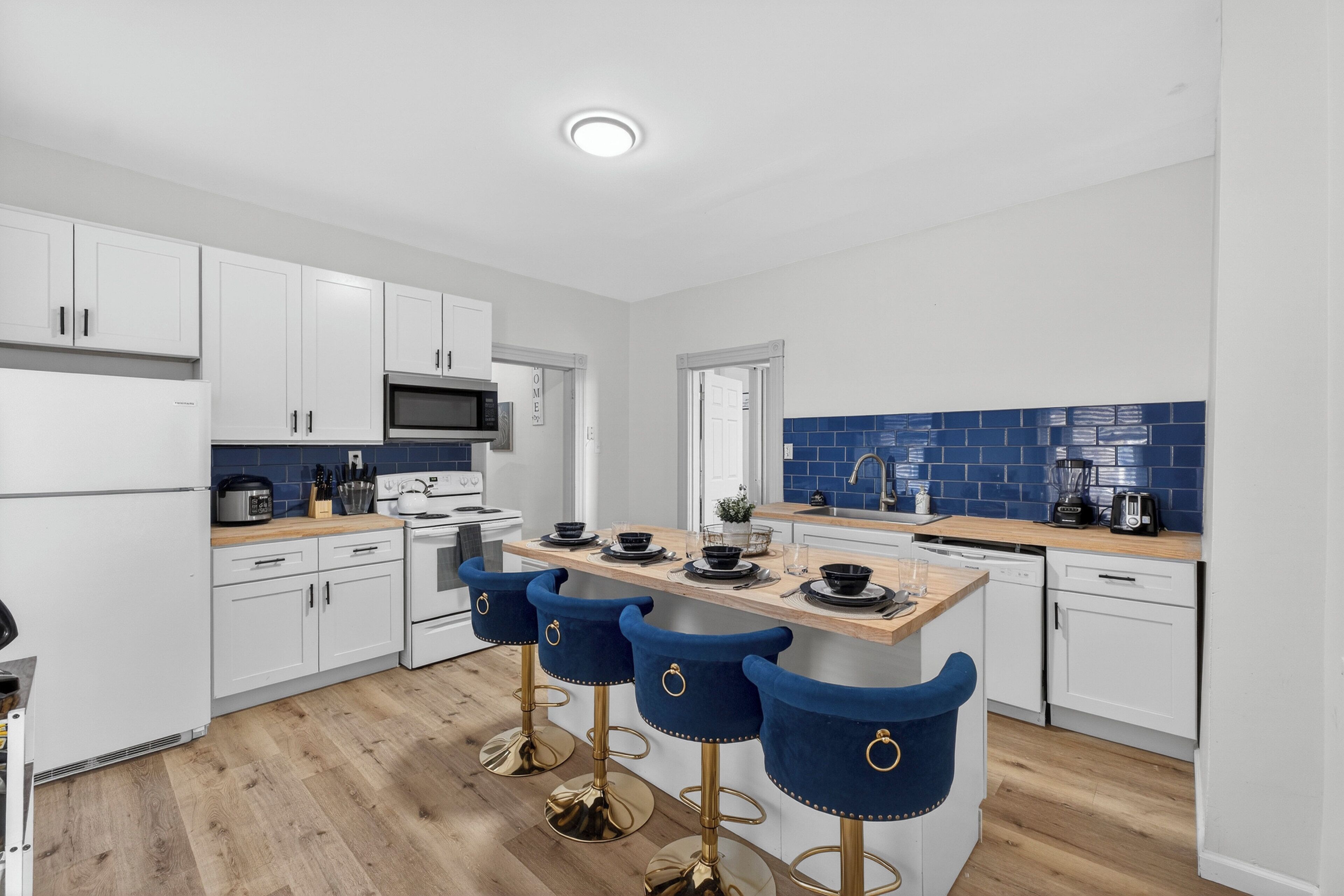 A modern kitchen with white cabinets, a central island with bar stools, a microwave, and a sink with blue tiles.