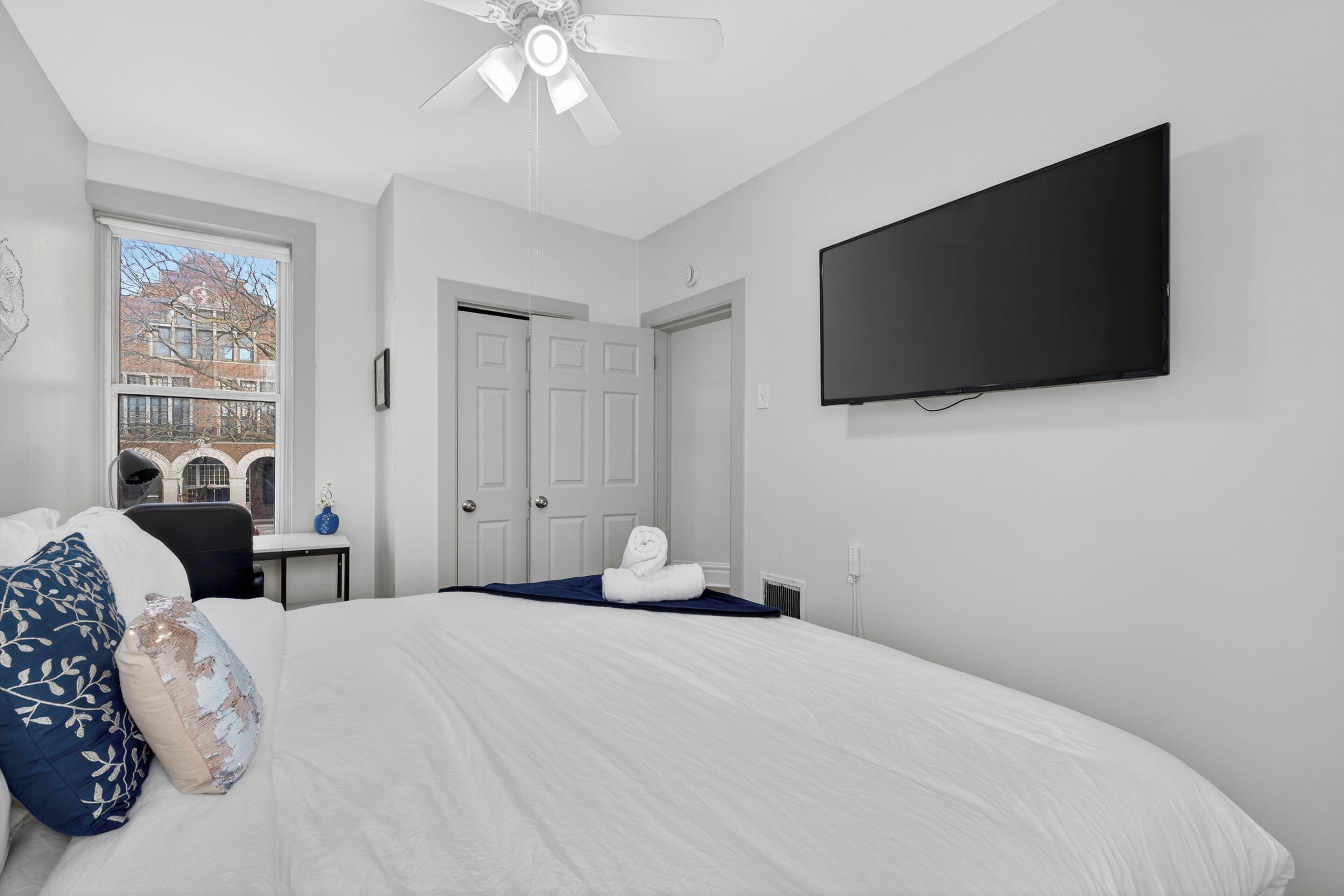 Une chambre à coucher comprenant un lit, un téléviseur à écran plat, un ventilateur de plafond et une fenêtre donnant sur des immeubles.