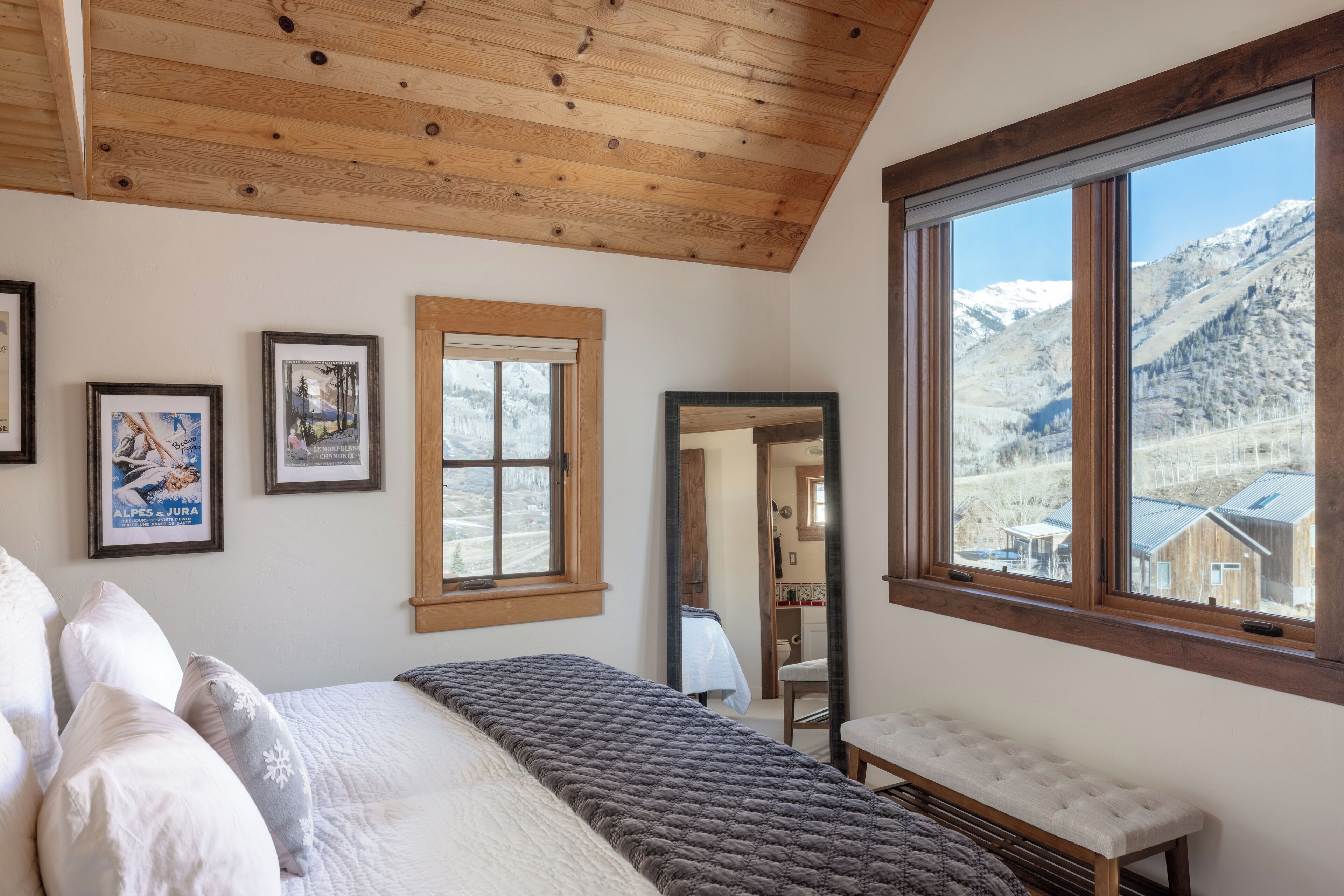 A bedroom with a wooden ceiling, a large window with a mountain view, a bed with pillows, and a bench.