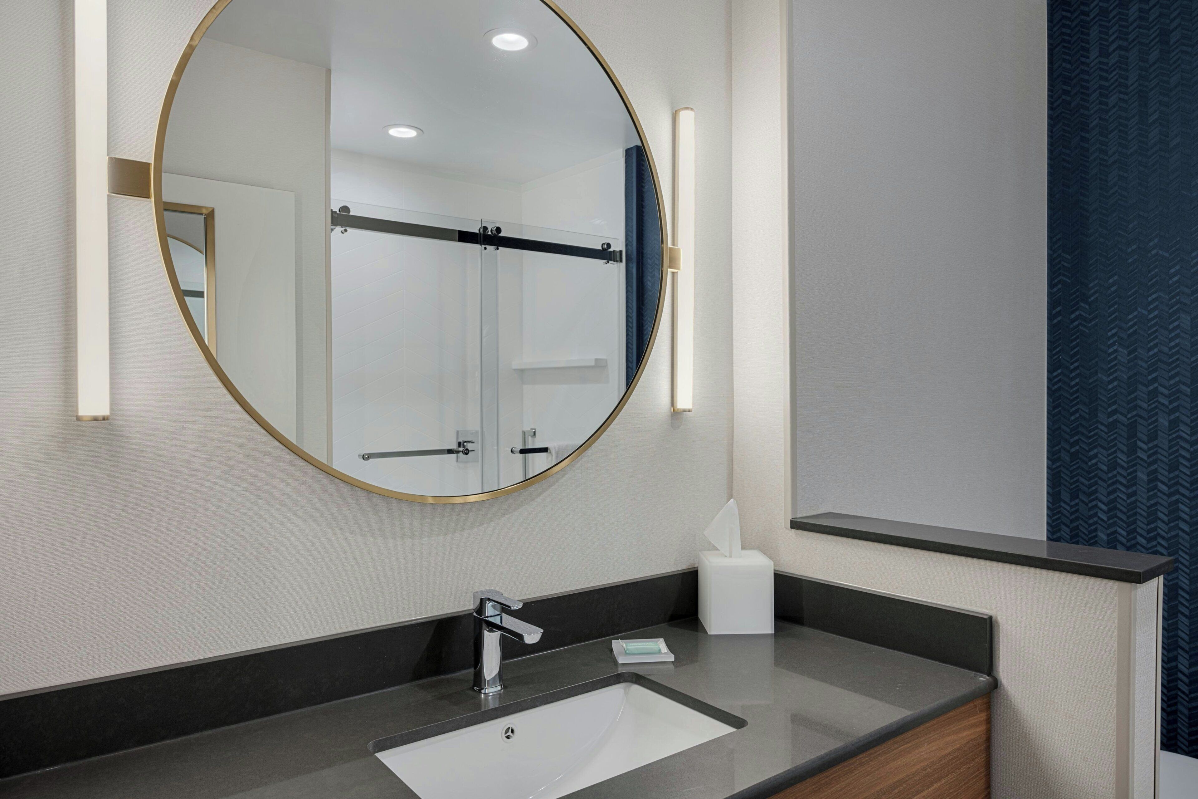 A modern bathroom with a round mirror, a sink, and a towel dispenser.