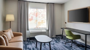 Une chambre d’hôtel avec un canapé, une petite table, un bureau avec une chaise, une télévision et une fenêtre avec des rideaux.