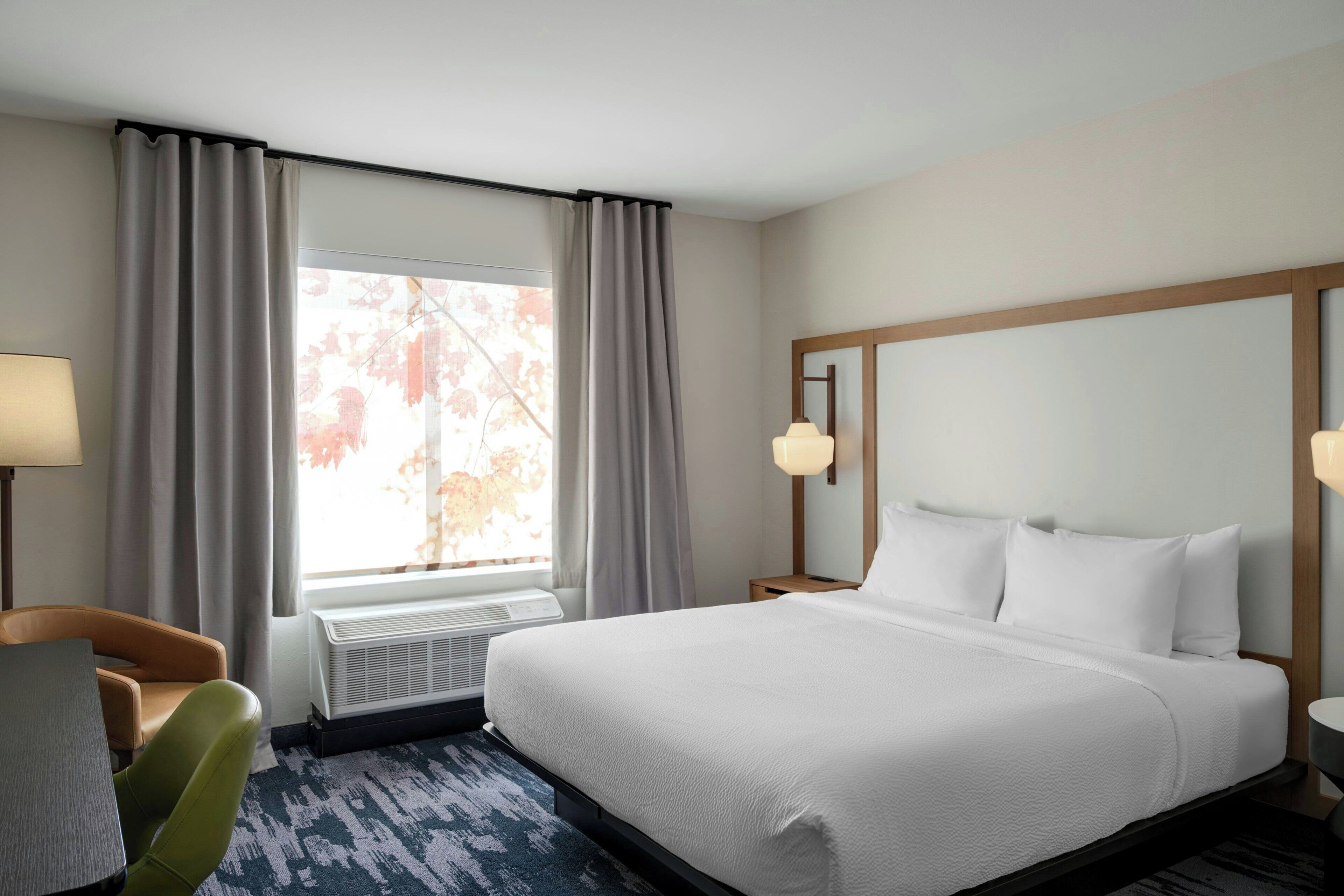 Habitación de hotel con cama, una silla, escritorio, lámpara y ventana con cortinas.