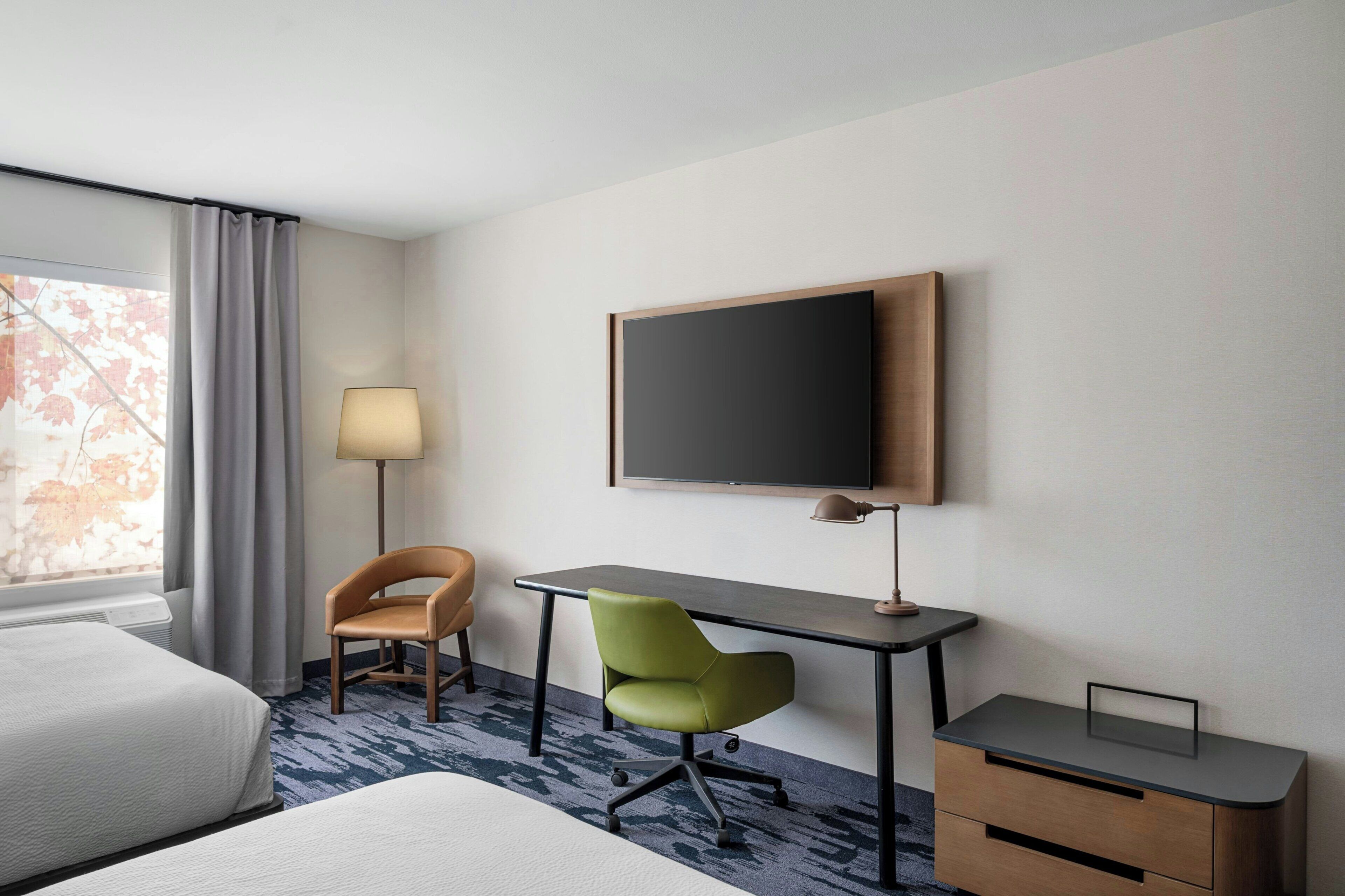 A hotel room with a bed, a desk, two chairs, a flat-screen TV, and a lamp.