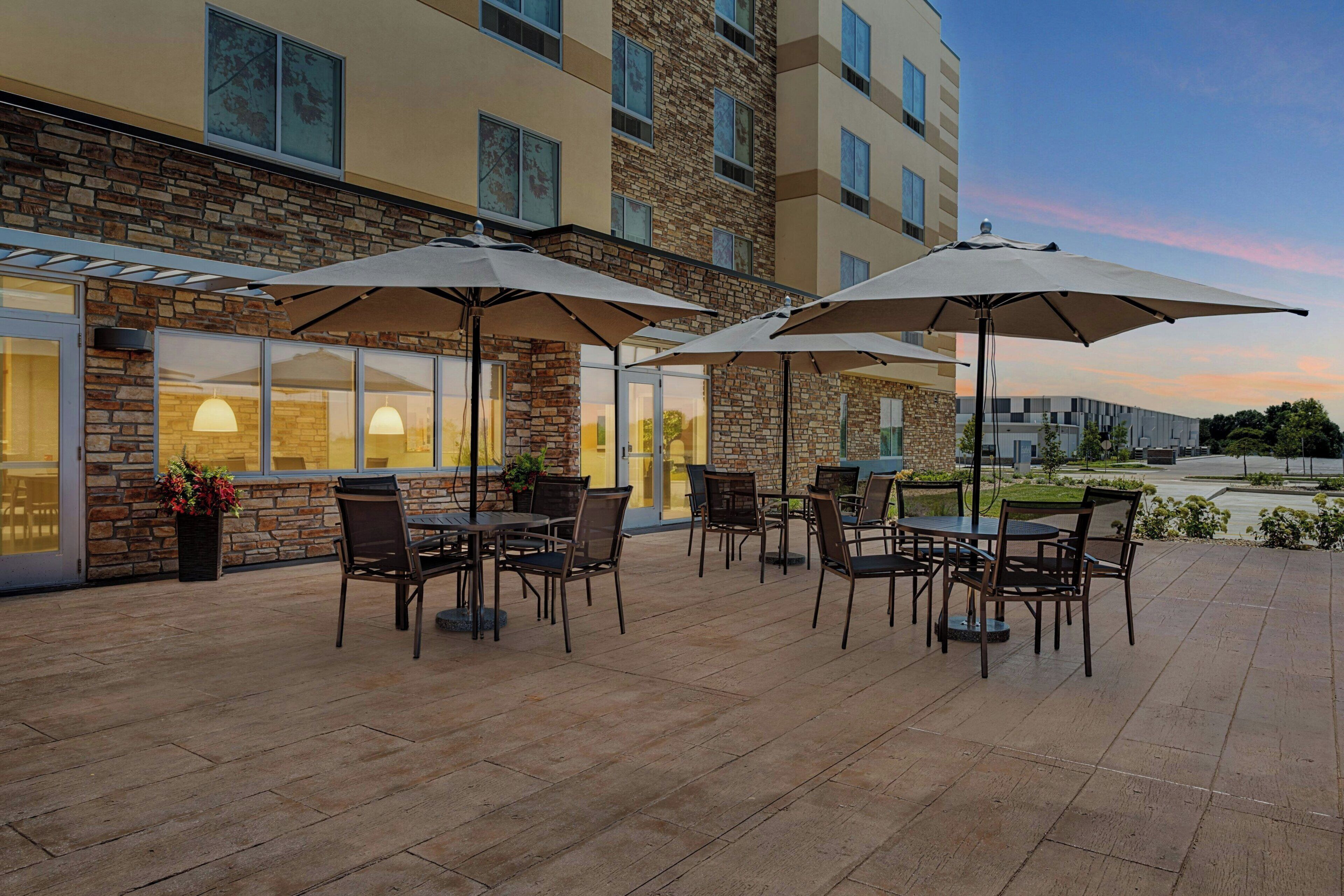 Un patio exterior con mesas y sillas, sombrillas y un edificio de fachada de piedra y ventanas.