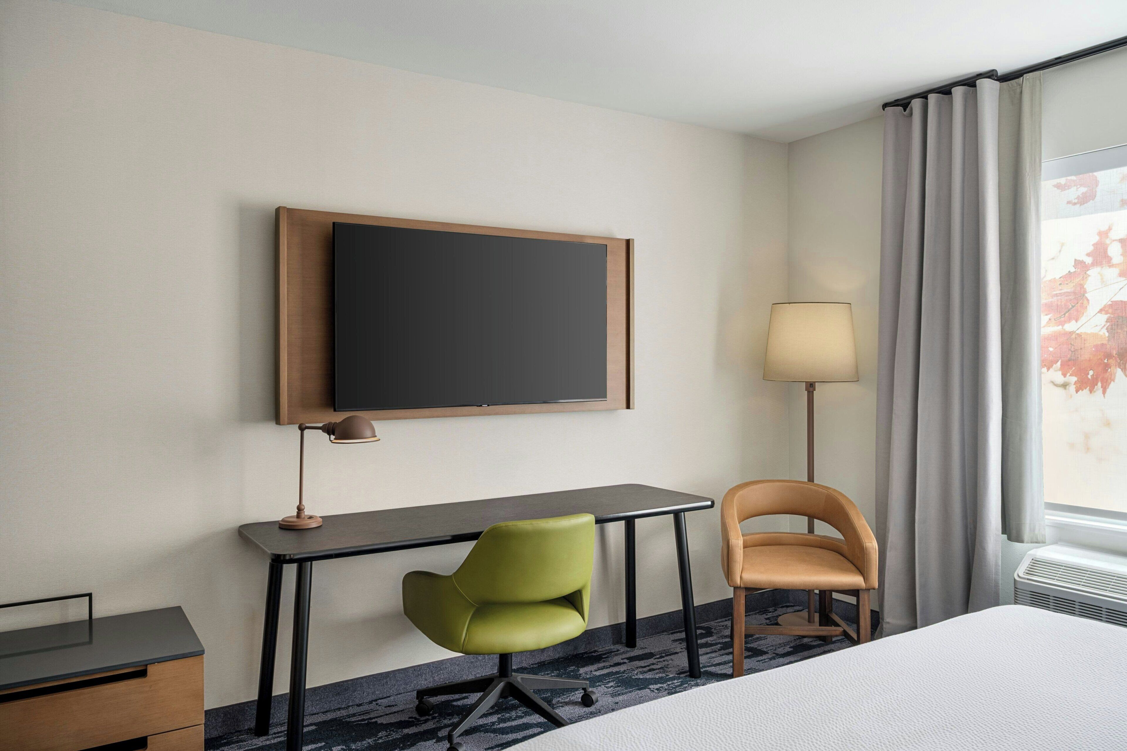 A hotel room with a bed, a desk, a green chair, a brown chair, a flat-screen TV, a lamp, and a window with curtains.