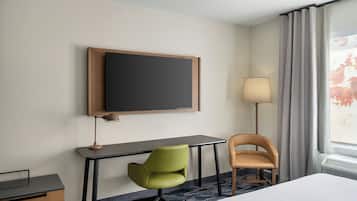 A hotel room with a bed, a desk, a green chair, a brown chair, a flat-screen TV, a lamp, and a window with curtains.