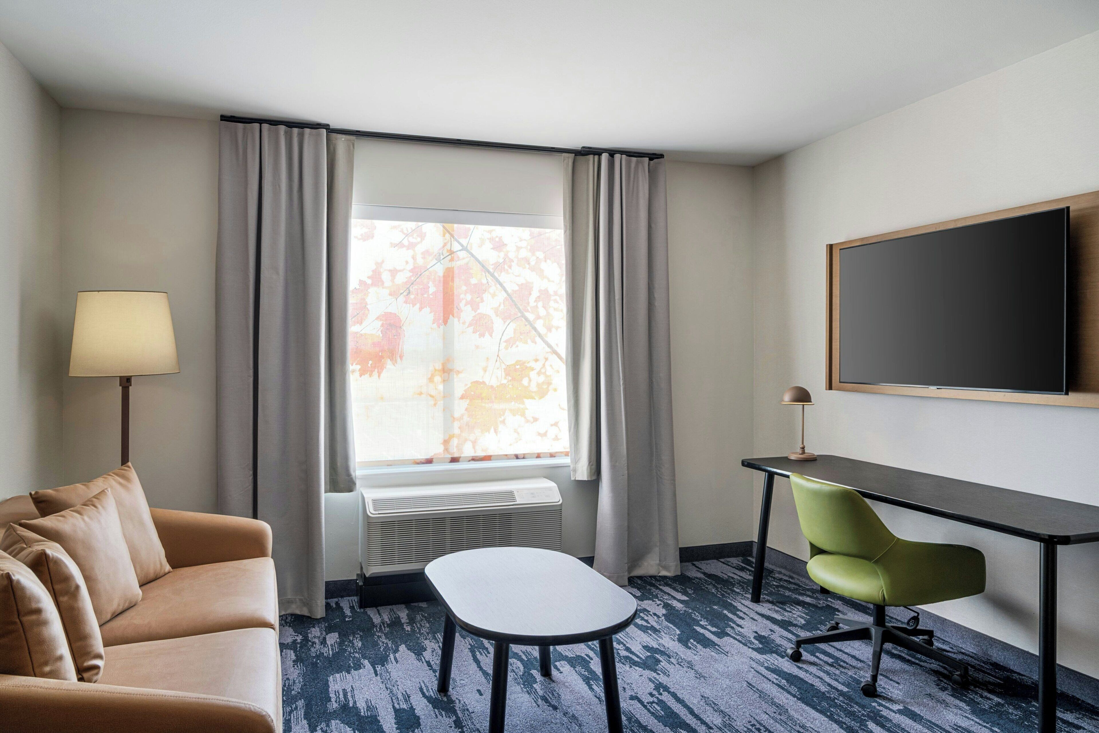 Une chambre d’hôtel comprenant un canapé, une petite table, un bureau avec une chaise, une grande fenêtre avec des rideaux et un téléviseur à écran plat fixé au mur.