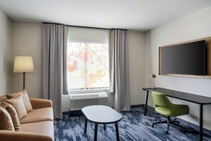 A hotel room with a sofa, a small table, a desk with a chair, a large window with curtains, and a flat-screen TV mounted on the wall. - Fairfield by Marriott Inn & Suites Mattoon (Mattoon)