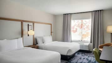 A hotel room with two beds, a desk, a chair, a window with curtains, and a wall-mounted lamp.