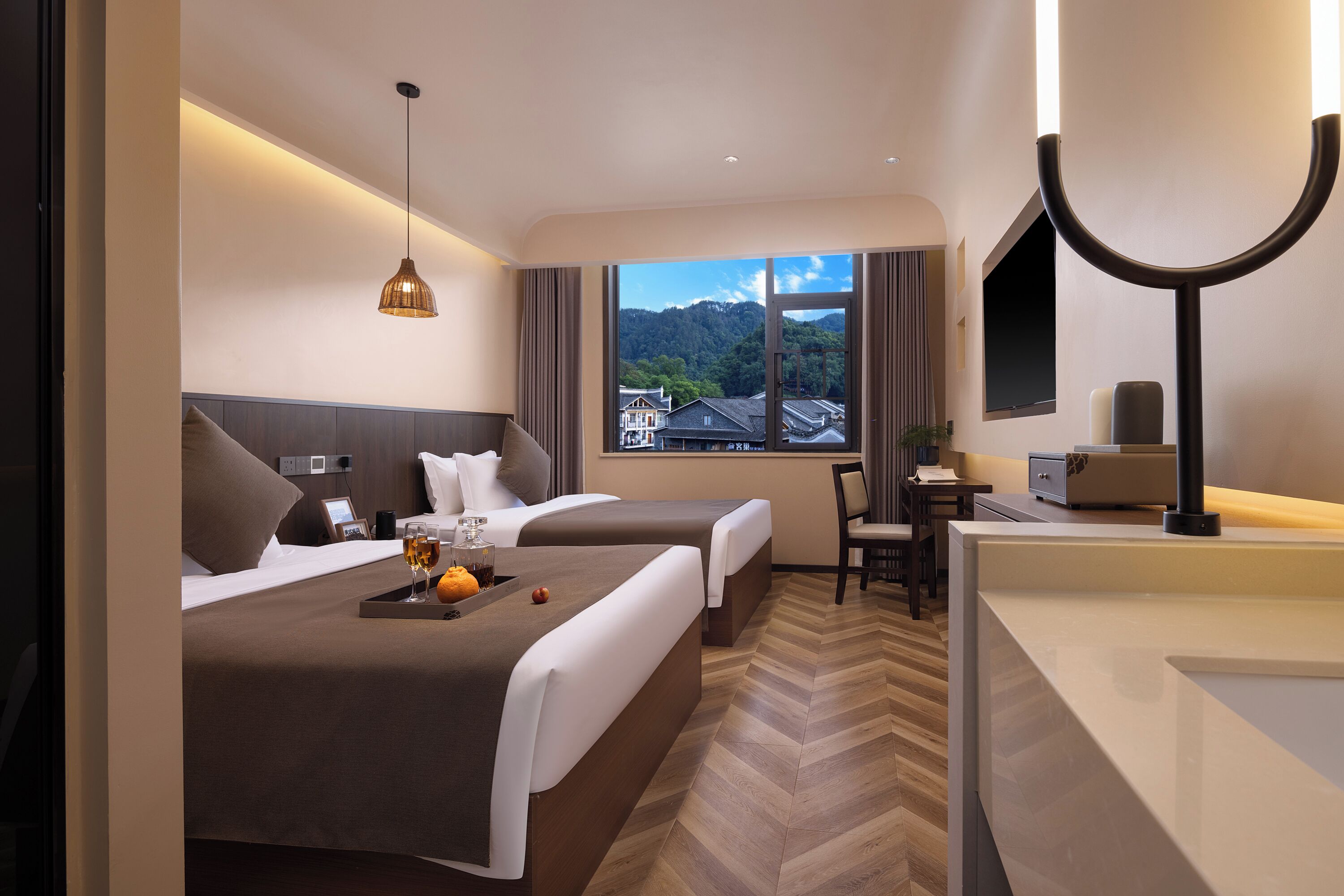 Une chambre d’hôtel avec deux lits, un bureau, une chaise, une grande fenêtre donnant sur des immeubles et des montagnes, et un luminaire suspendu moderne.