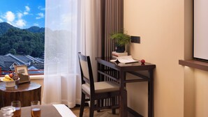 A hotel room with a wooden chair, a small table with a plant, a framed picture, and a view of a traditional building through a window. - Fenghuang Story Hotel (Xiangxi)