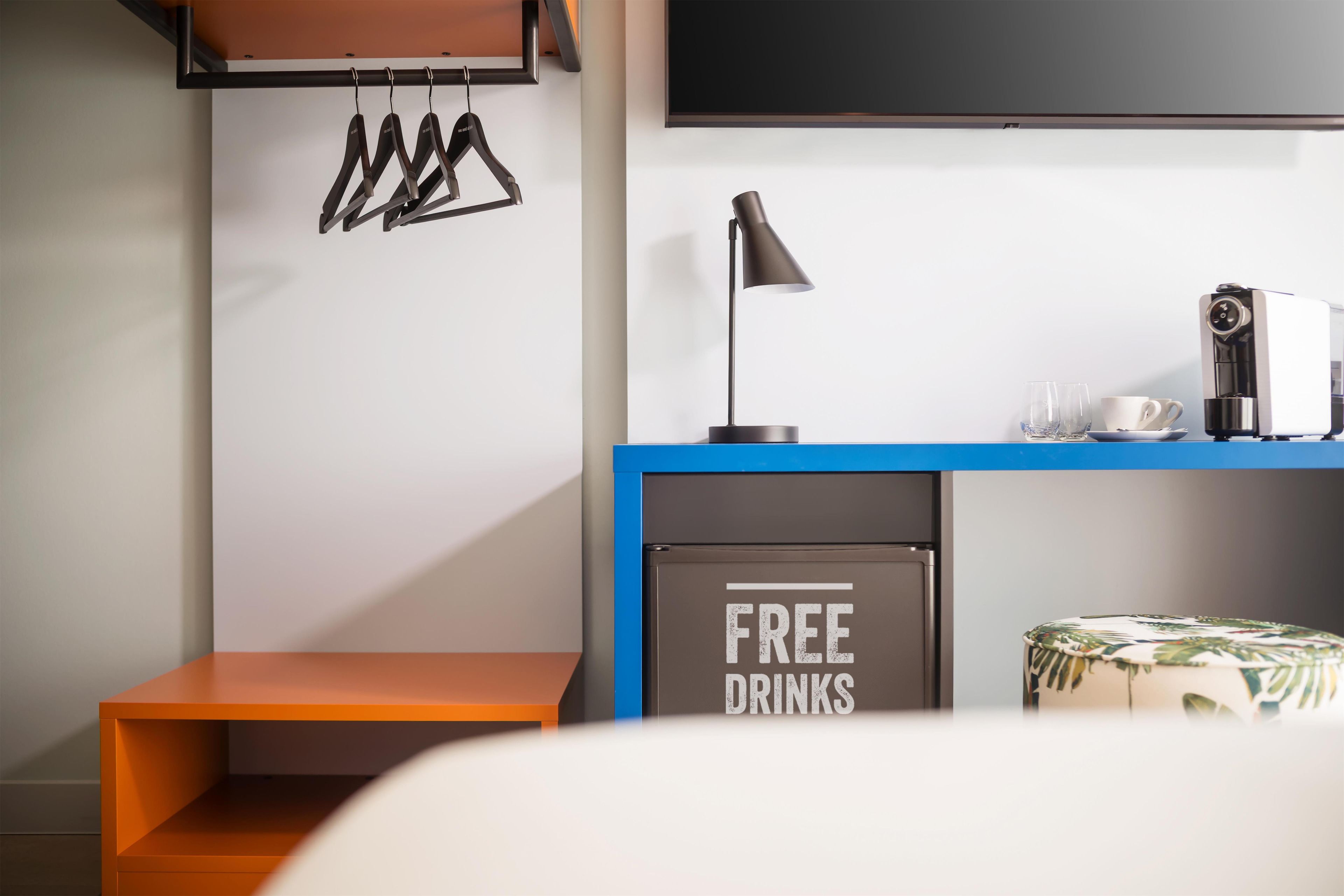 A modern room with a blue desk, a coffee machine, a sign offering free drinks, a hanging coat rack, and a television mounted on the wall.