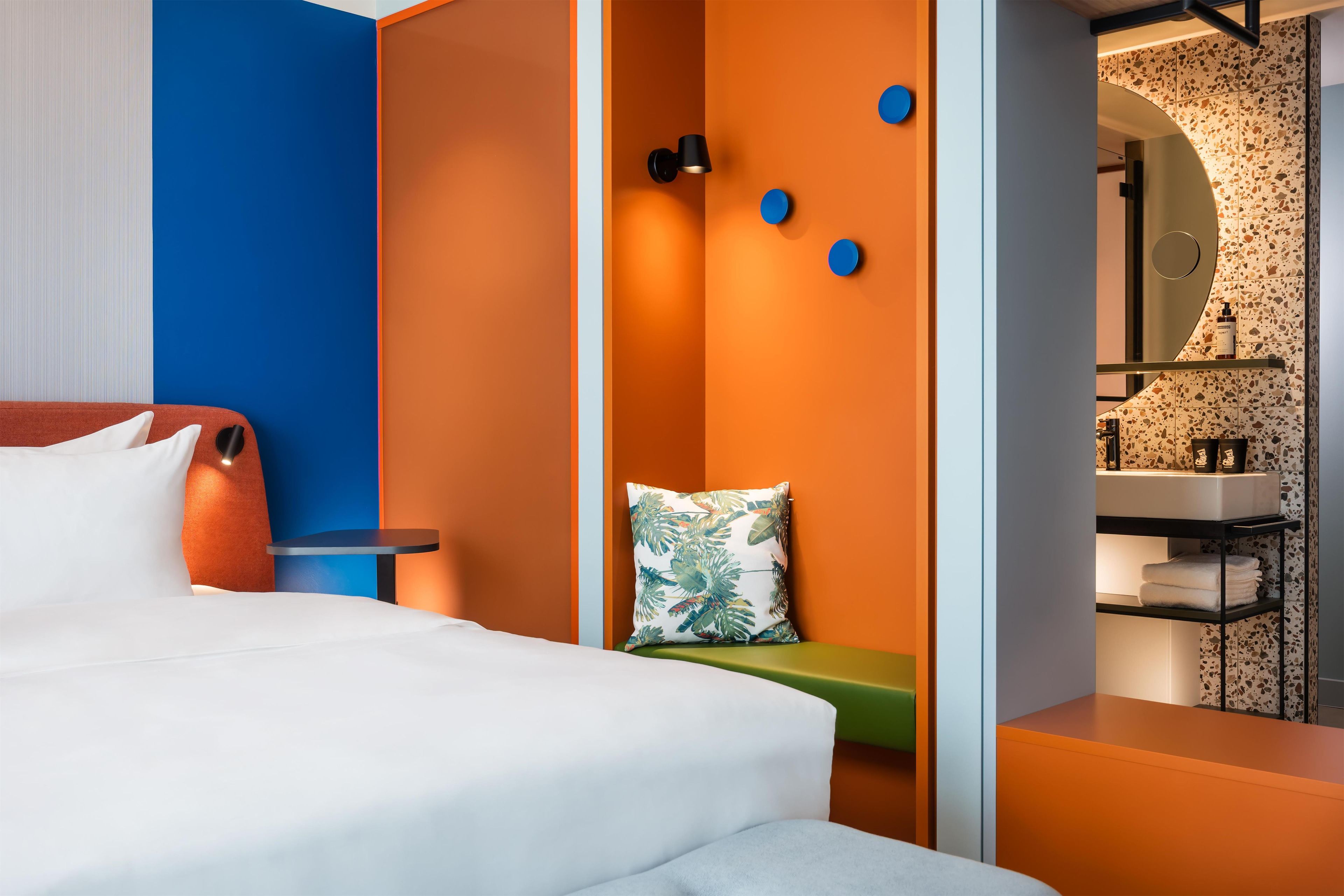 Ein modernes Hotelzimmer mit Bett, kleinem Tisch, Bank und einem Badezimmer mit Waschbecken und Spiegel.