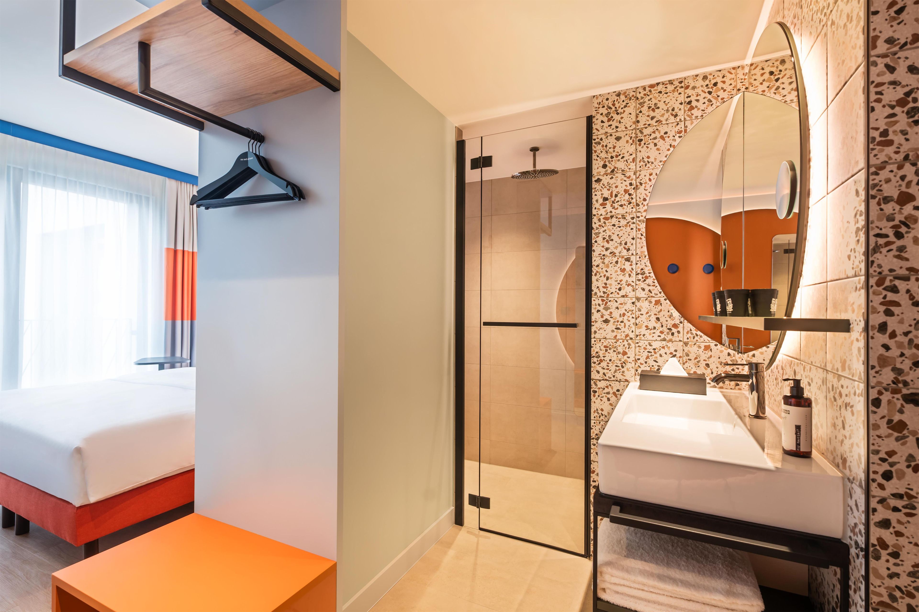 Ein Hotelzimmer mit einem Bett, einem kleinen Tisch, einer Duschkabine mit Glastür sowie einem Badezimmer mit Waschbecken und Spiegel.