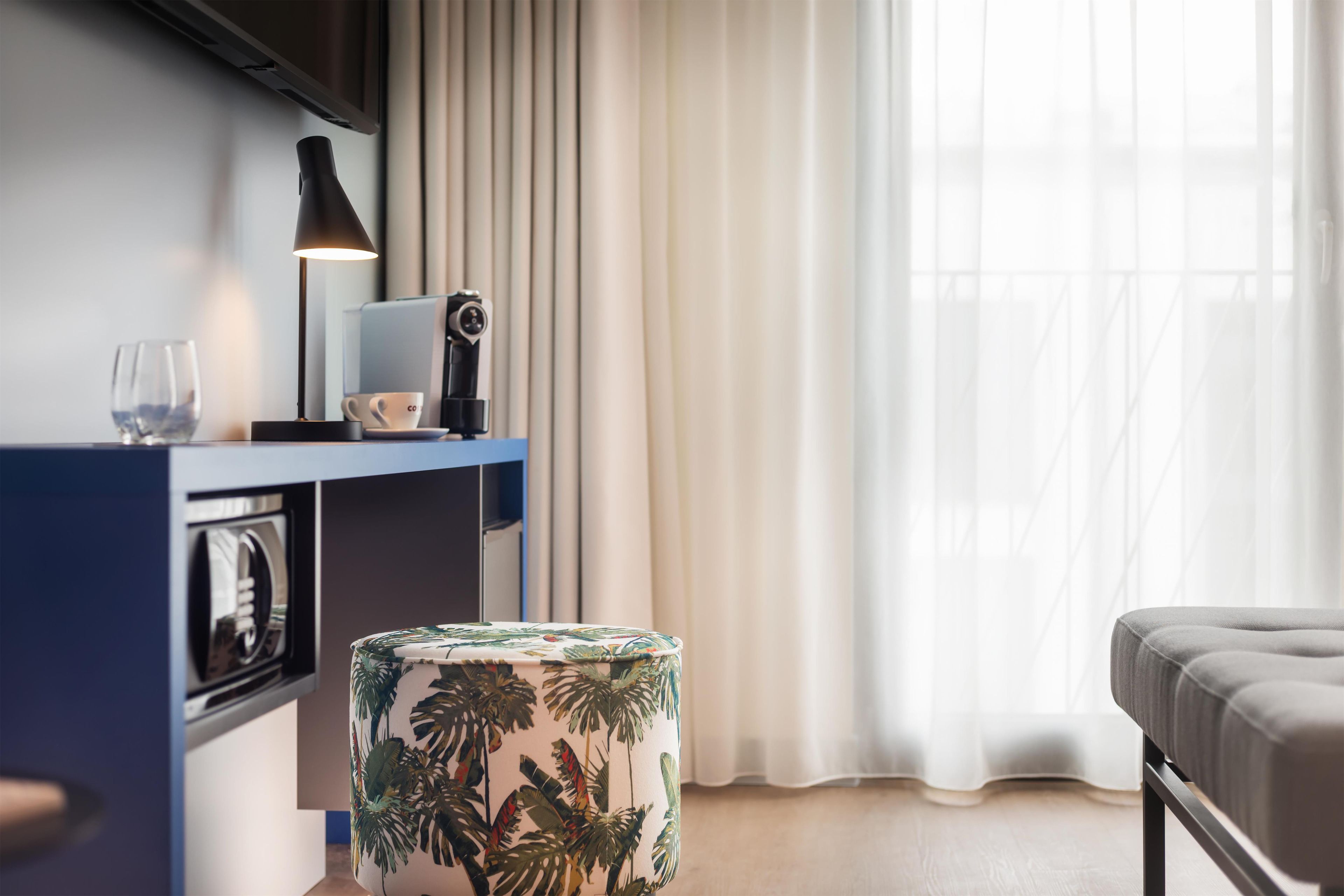 Ein modernes Hotelzimmer mit Schreibtisch, einem Laptop, einer Kamera, einer Tasse, einem Glas und einem osmanischen Hocker im tropischen Stil.