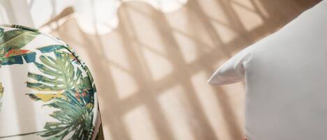 A room with a window casting shadows on a white pillow and a fabric with tropical leaf patterns.