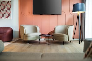 A modern living room with two armchairs, a small round table, a floor lamp, and a flat-screen TV mounted on a wall with a geometric patterned artwork. - Me and All Hotel Leipzig, by Hyatt (Leipzig)