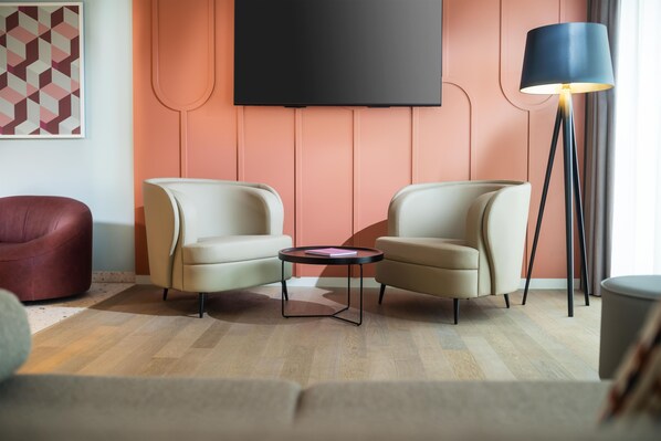 Una sala de estar moderna con dos sillones, una mesita redonda, una lámpara de pie y un televisor de pantalla plana montado en la pared, junto con una obra de arte con diseño geométrico.