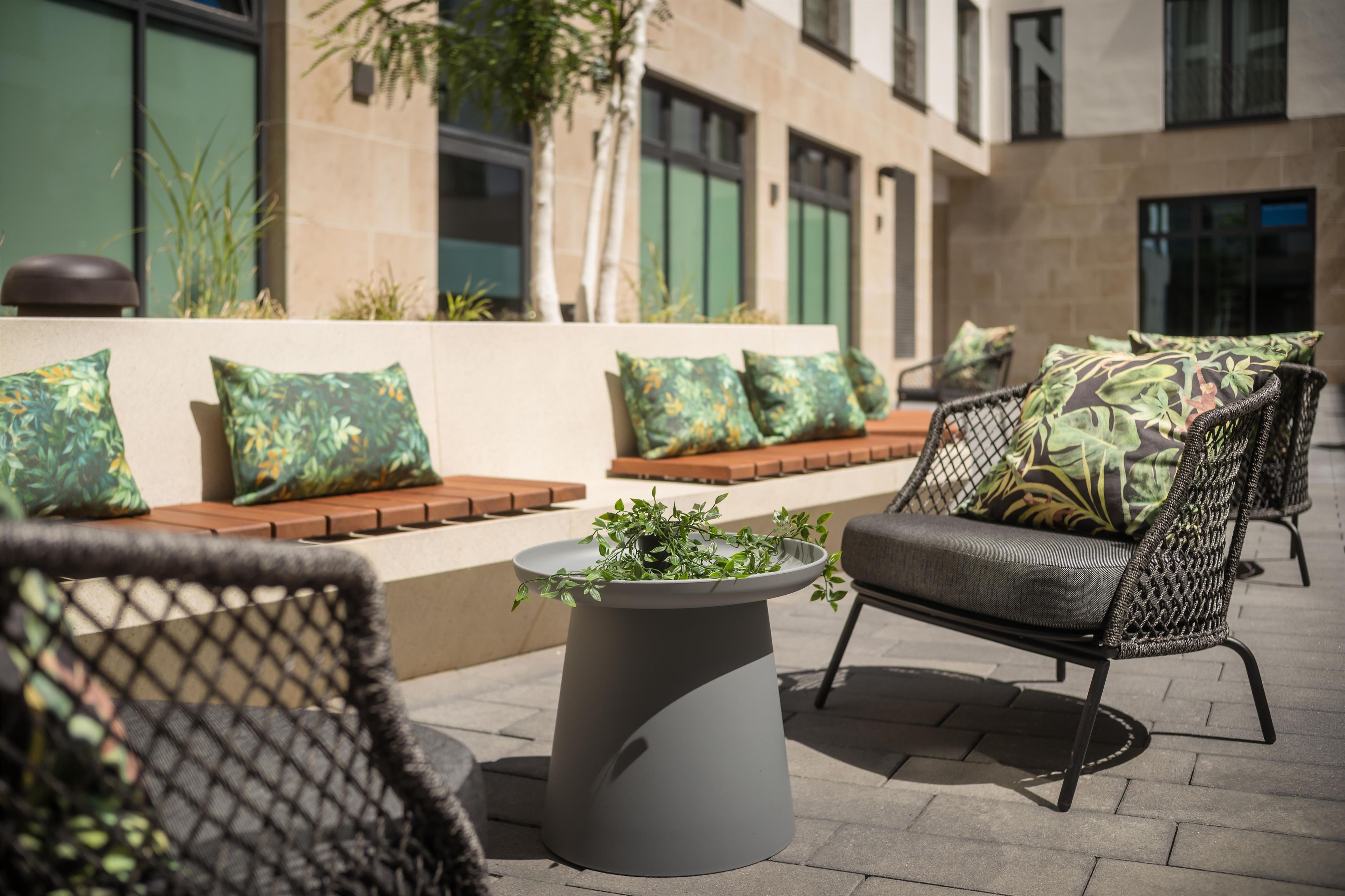 An outdoor seating area with wicker chairs and cushions, a central planter with greenery, and a building with large windows in the background.